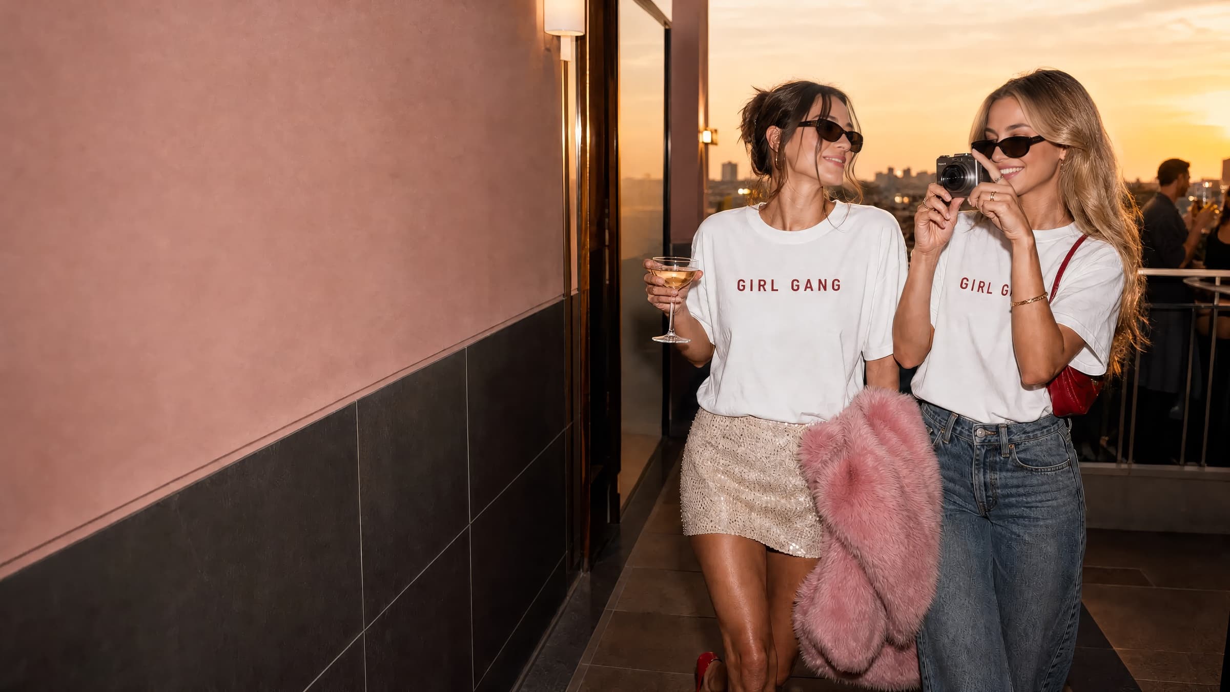 Two friends in matching Girl Gang tees on a rooftop at sunset, one holding a martini and one a camera